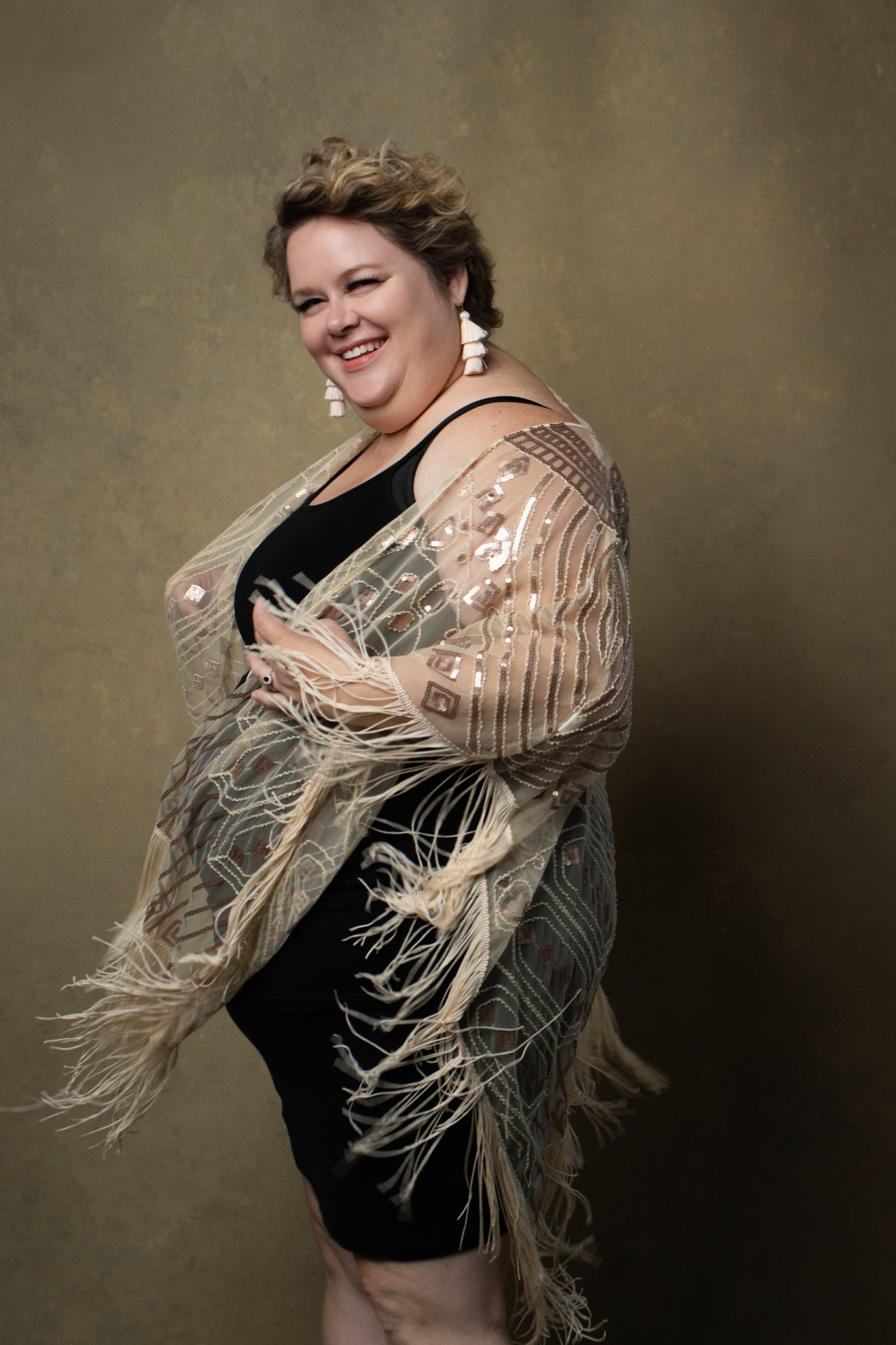A woman in her fifties with short auburn hair and tasselled earrings, laughing, in a black dress under a gold sequinned fringe wrap.