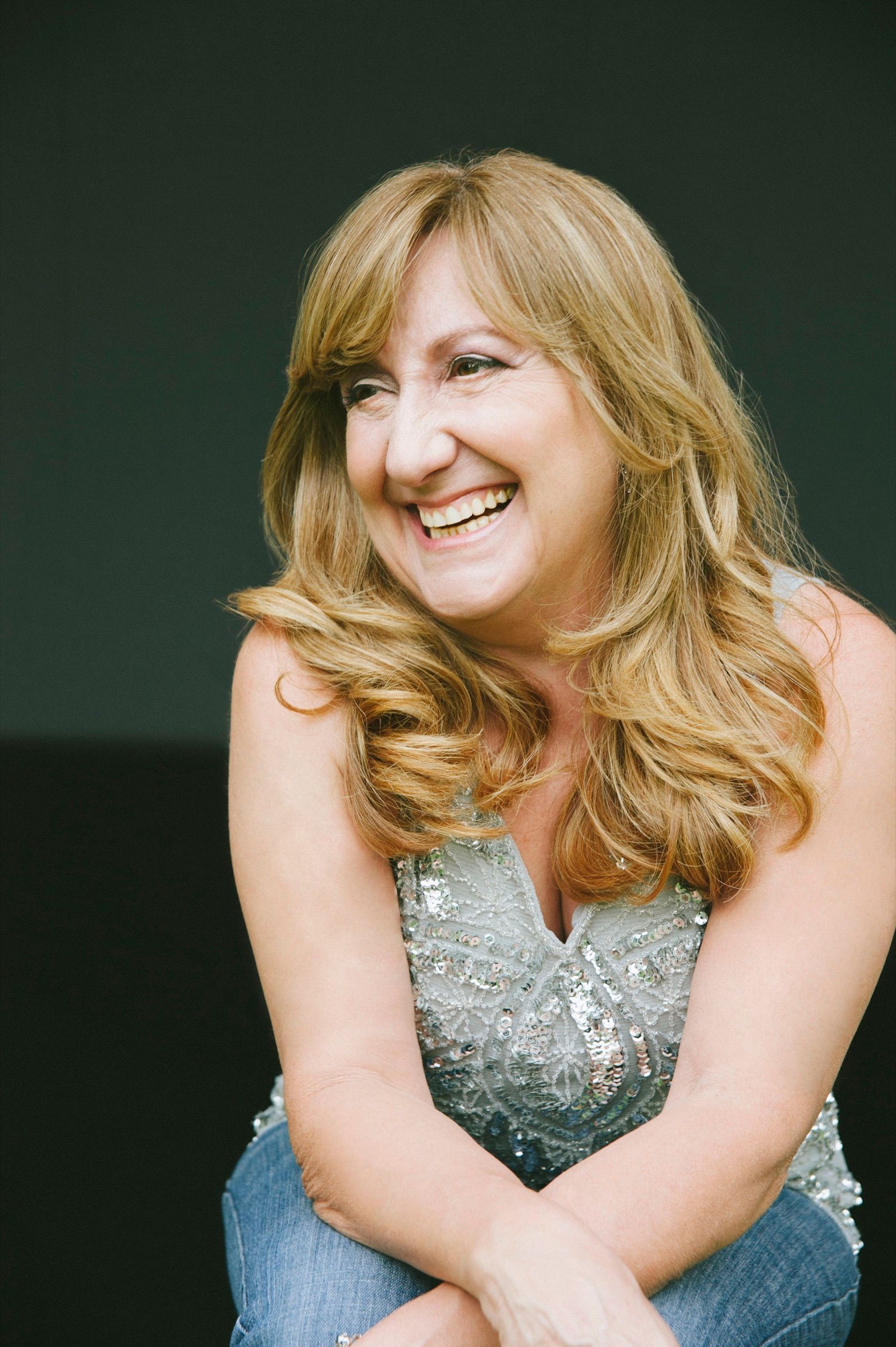 A woman of fifty with long blonde hair, laughing and looking away, seated in a silver sequin top and jeans.