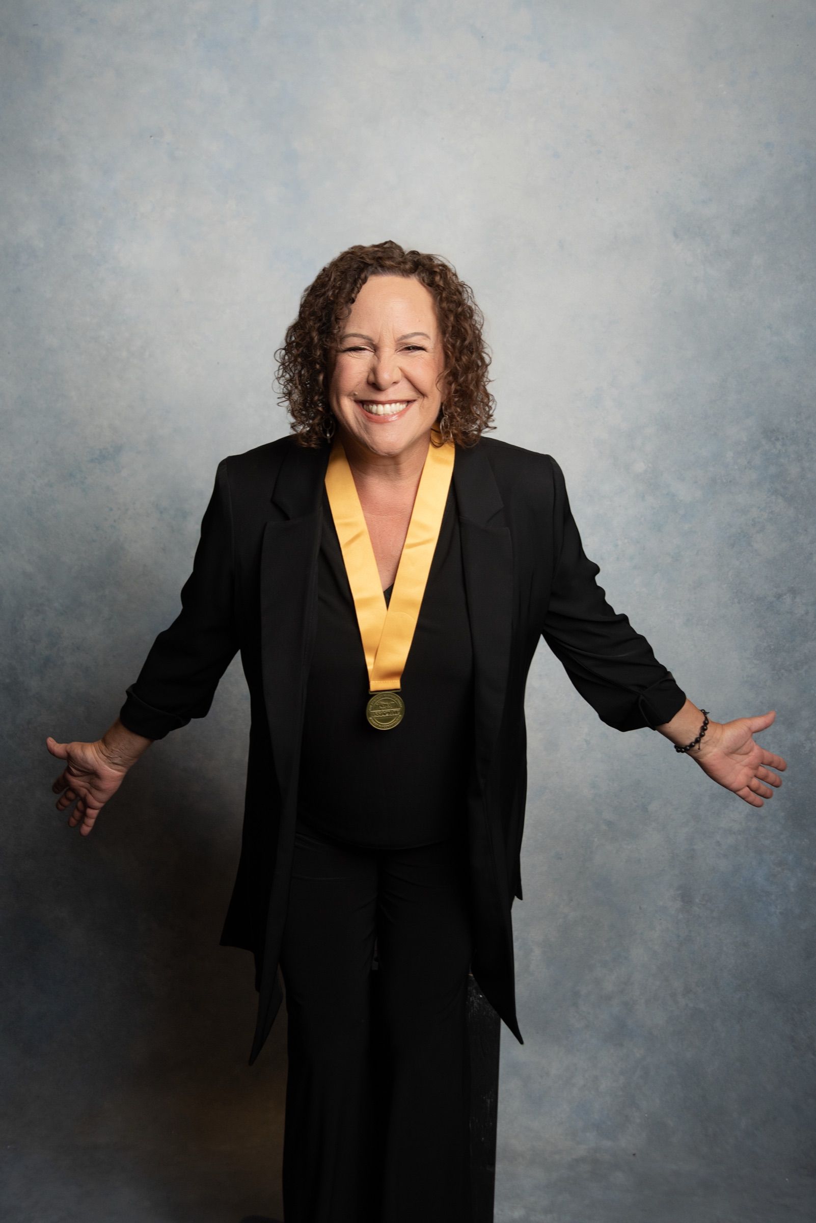 A woman in her sixties with dark curls, laughing with her arms open, a gold medal over her black suit.
