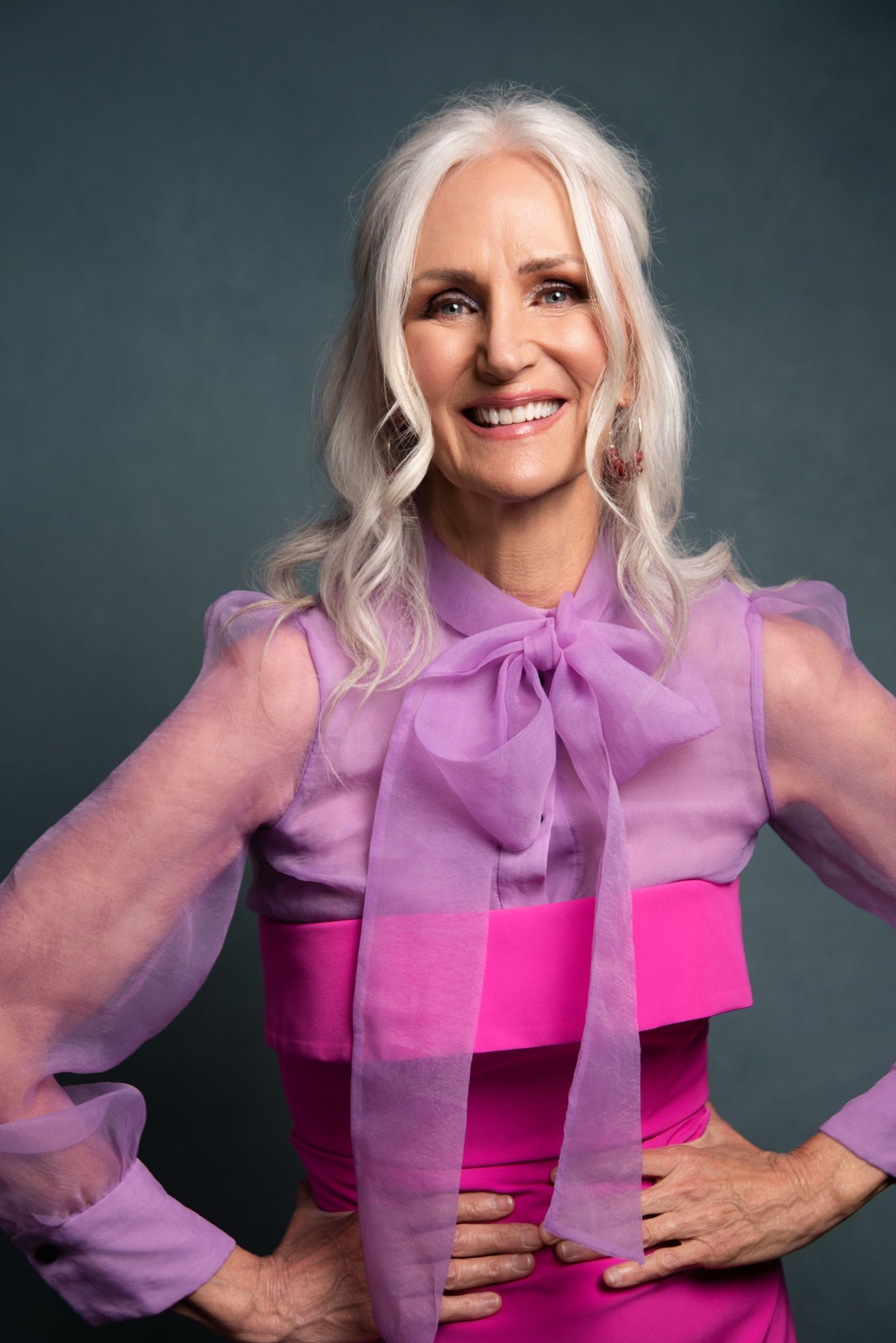 A woman in her sixties with long platinum hair, smiling, hands on hips in a lilac pussy-bow blouse over magenta.