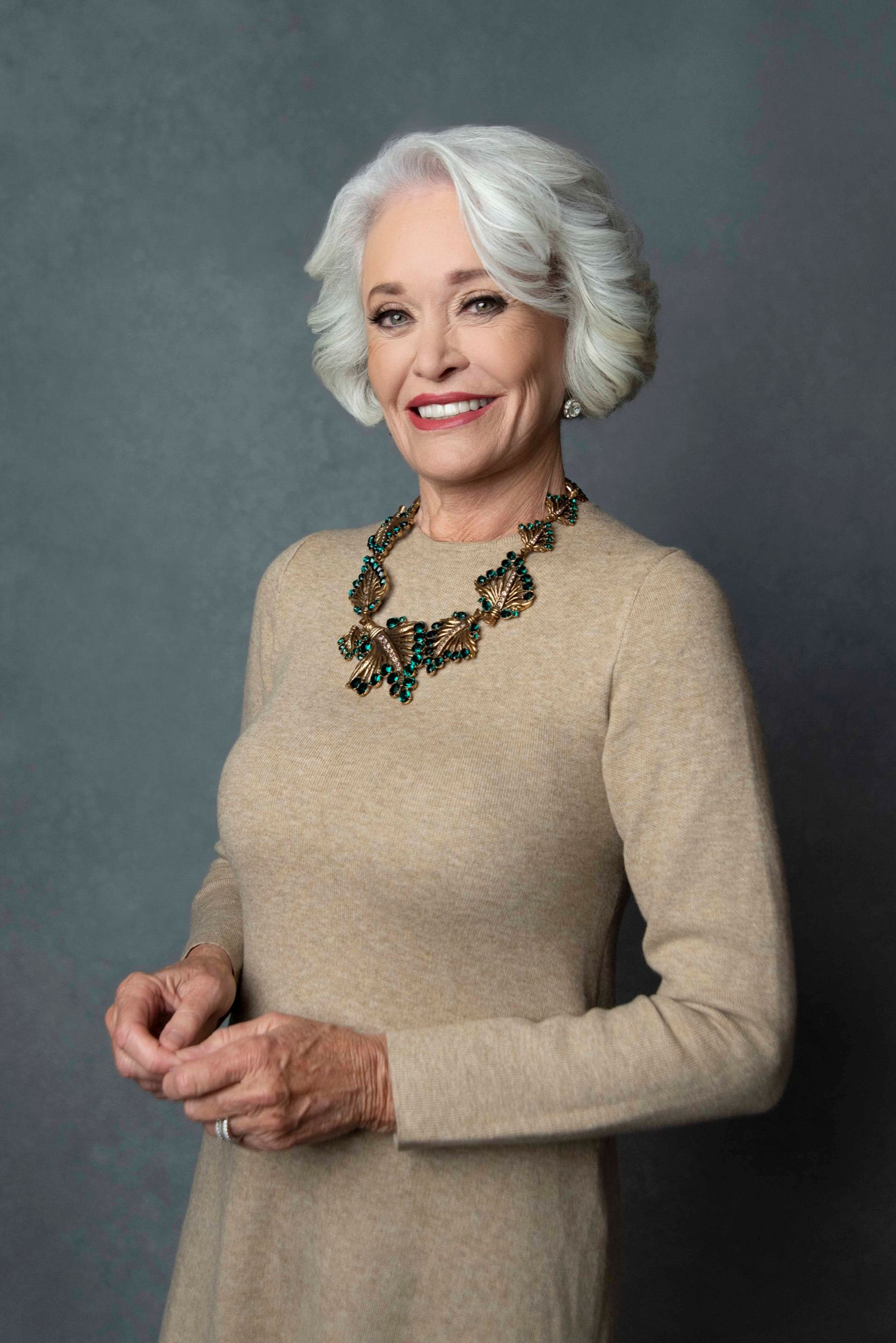 A woman around seventy with a silver bob and a red lip, hands clasped, in a tan knit and a gold and emerald necklace.